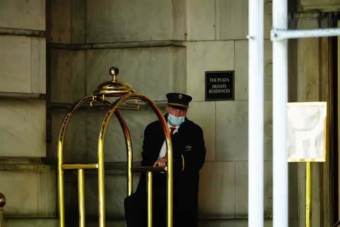 A hotel doorman wearing a mask stands outside the hotel next to a luggage cart